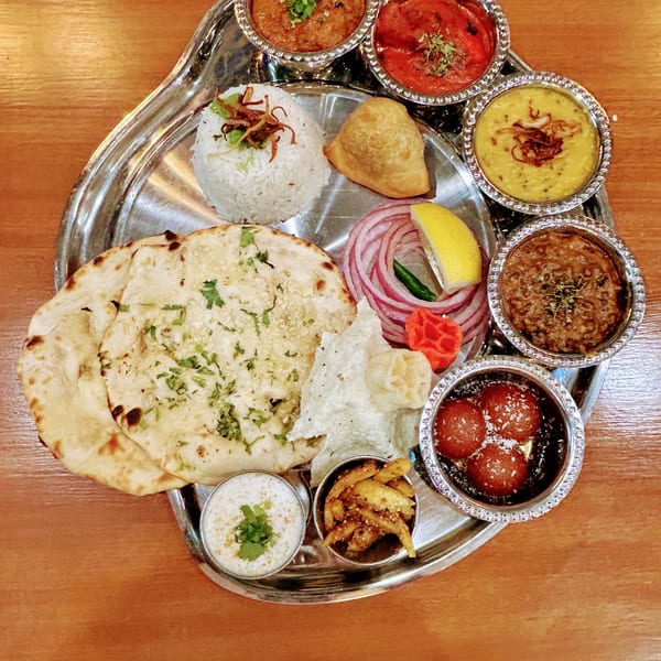 A silver platter with various Indian dishes including naan, rice, curries, samosa, and gulab jamun.